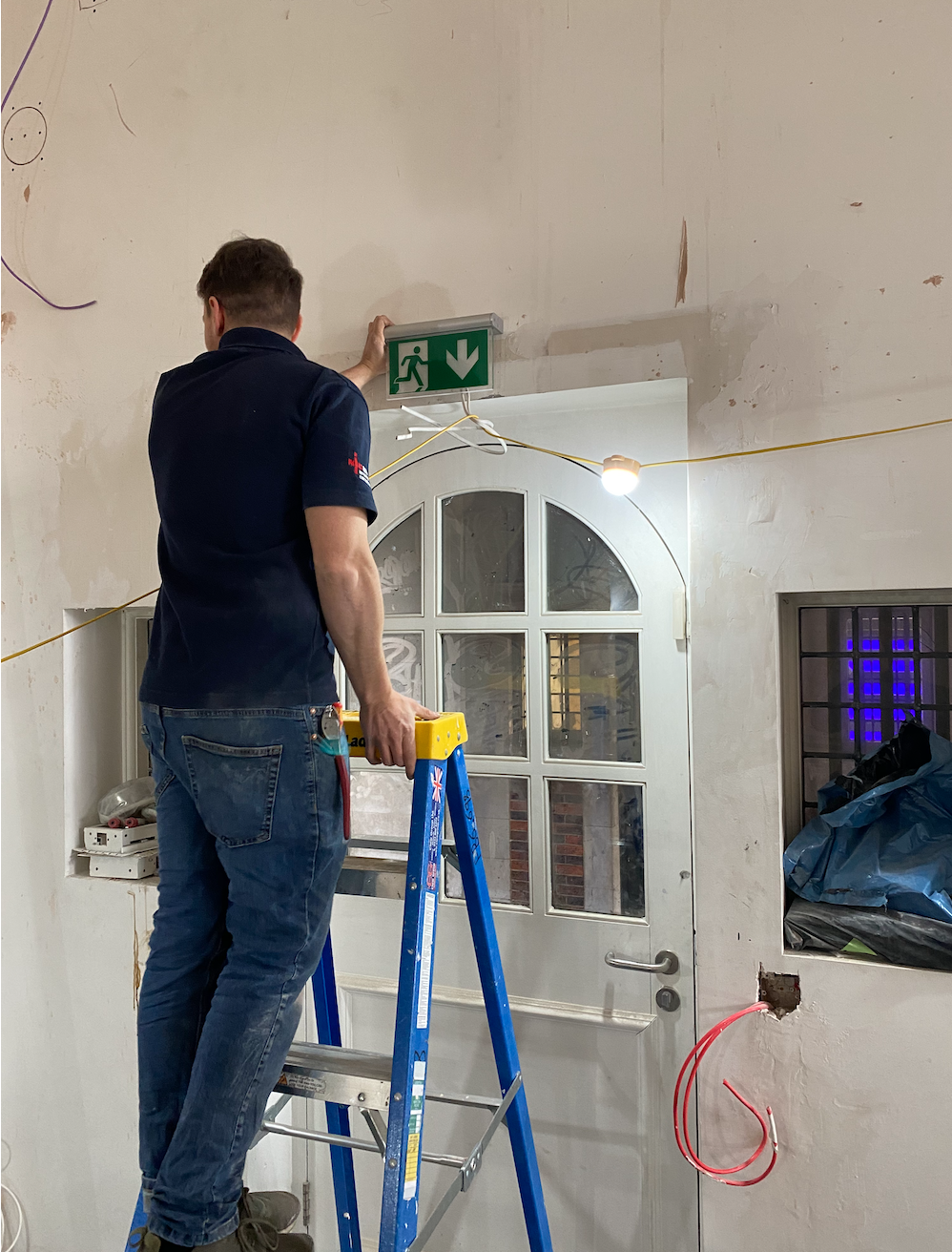 Electrician on a step ladder fitting an illuminated emergency exit sign above a doorway